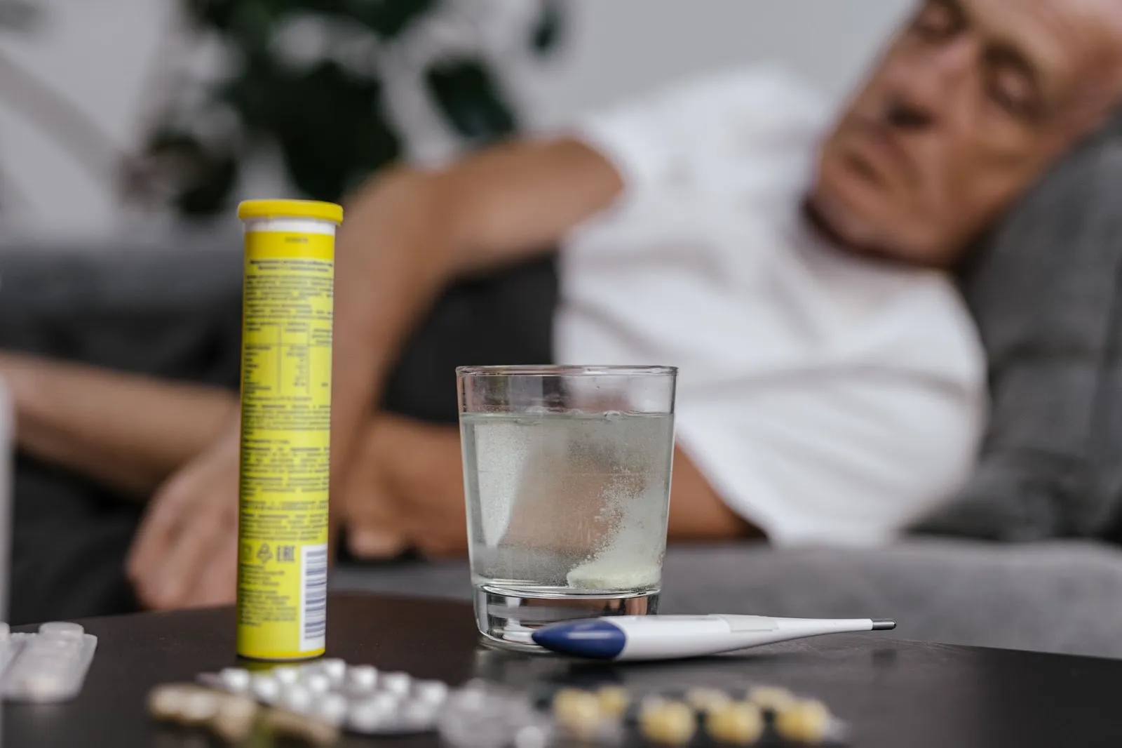 Man sitting on couch looking at medicine, glass of water, and thermometer sitting on desk
